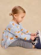 A toddler girl sitting on a beige studio floor, wearing a light blue long-sleeve top with an orange and white dog print, navy and white striped leggings, and navy sneakers.