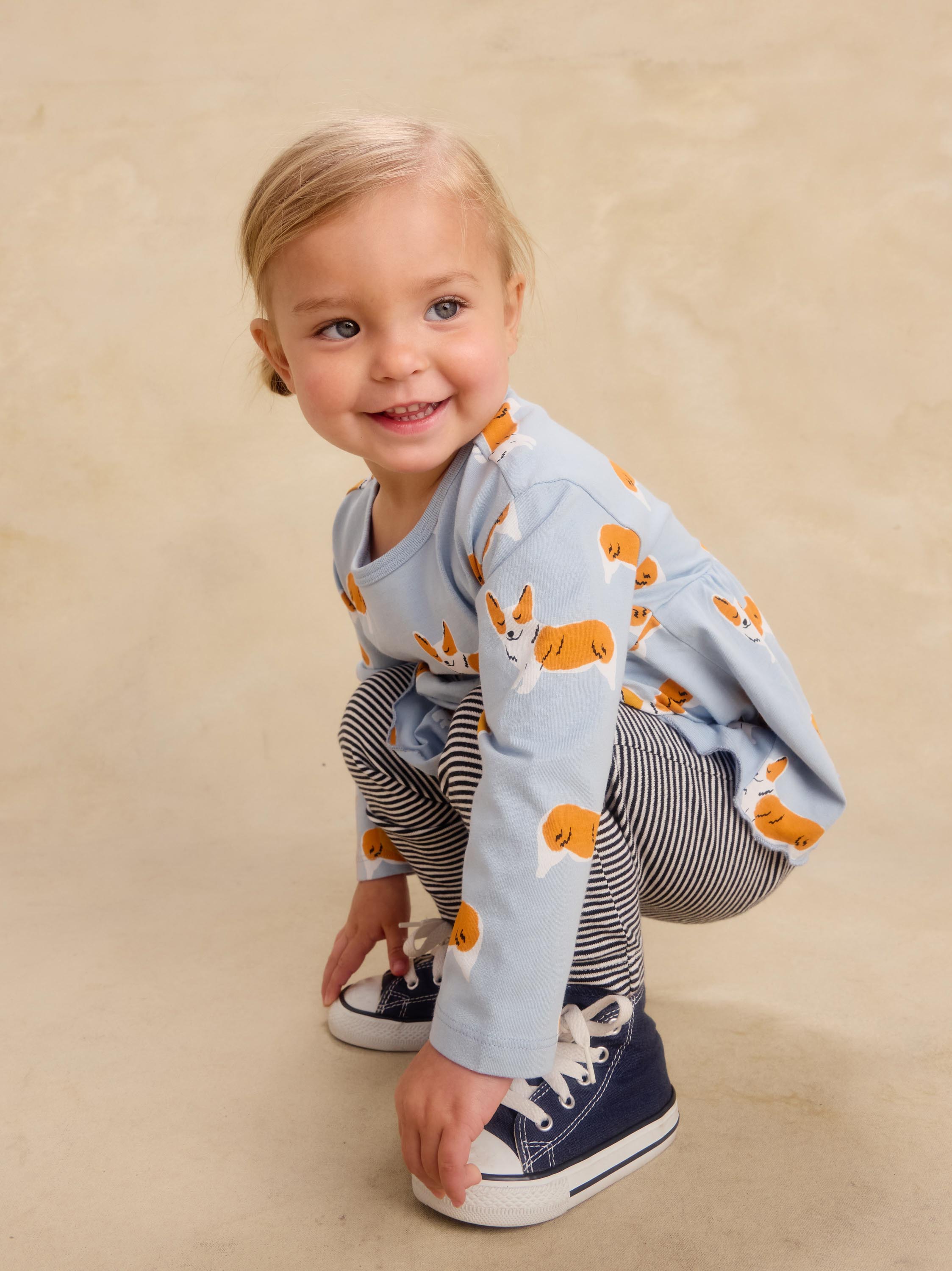 A toddler girl crouching and smiling while wearing a light blue long-sleeve top with an orange and white dog print, navy and white striped pants, and navy sneakers, set against a warm beige studio backdrop.