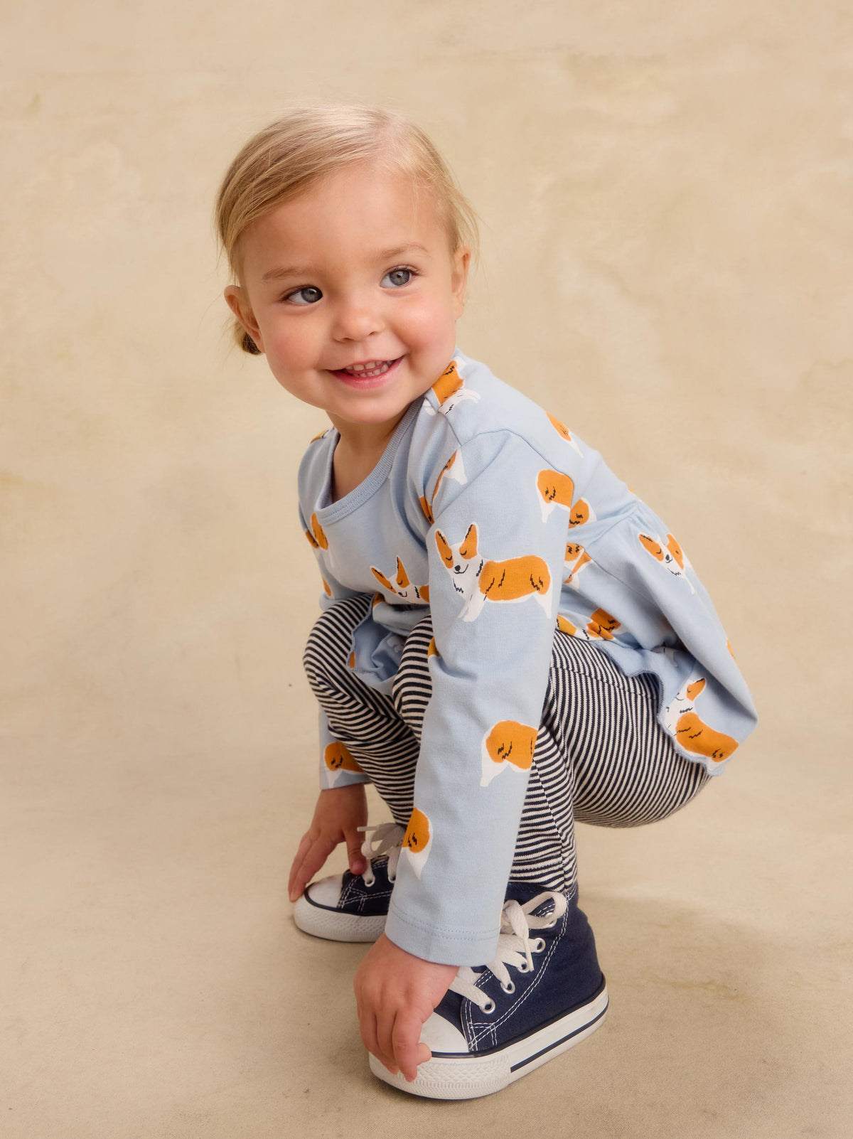 A toddler girl crouching and smiling while wearing a light blue long-sleeve top with an orange and white dog print, navy and white striped pants, and navy sneakers, set against a warm beige studio backdrop.