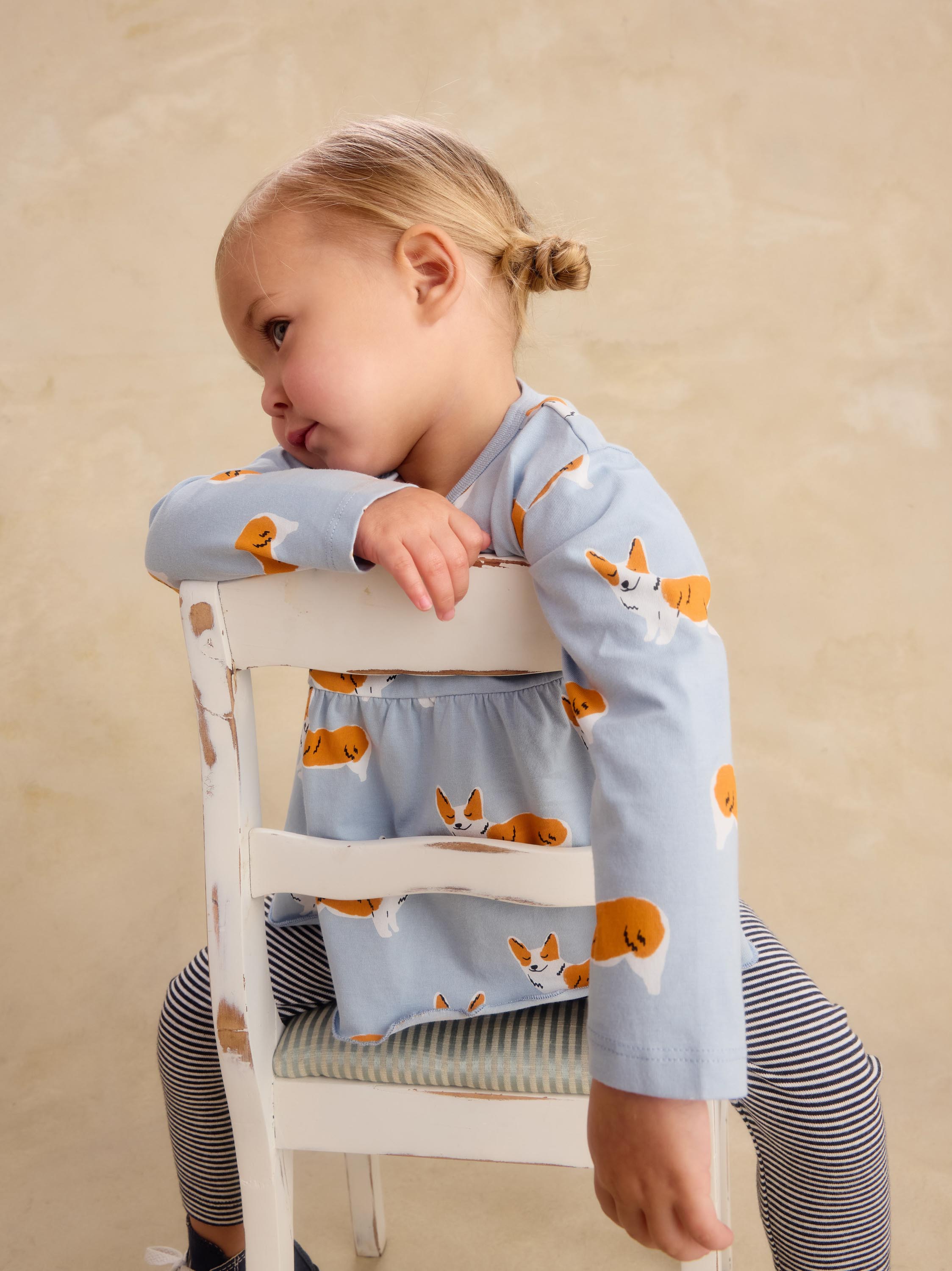 A young child wearing a light blue long-sleeve top and striped leggings sits on a white wooden chair, leaning on the backrest against a warm beige background.