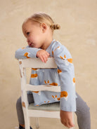 A young child wearing a light blue long-sleeve top and striped leggings sits on a white wooden chair, leaning on the backrest against a warm beige background.