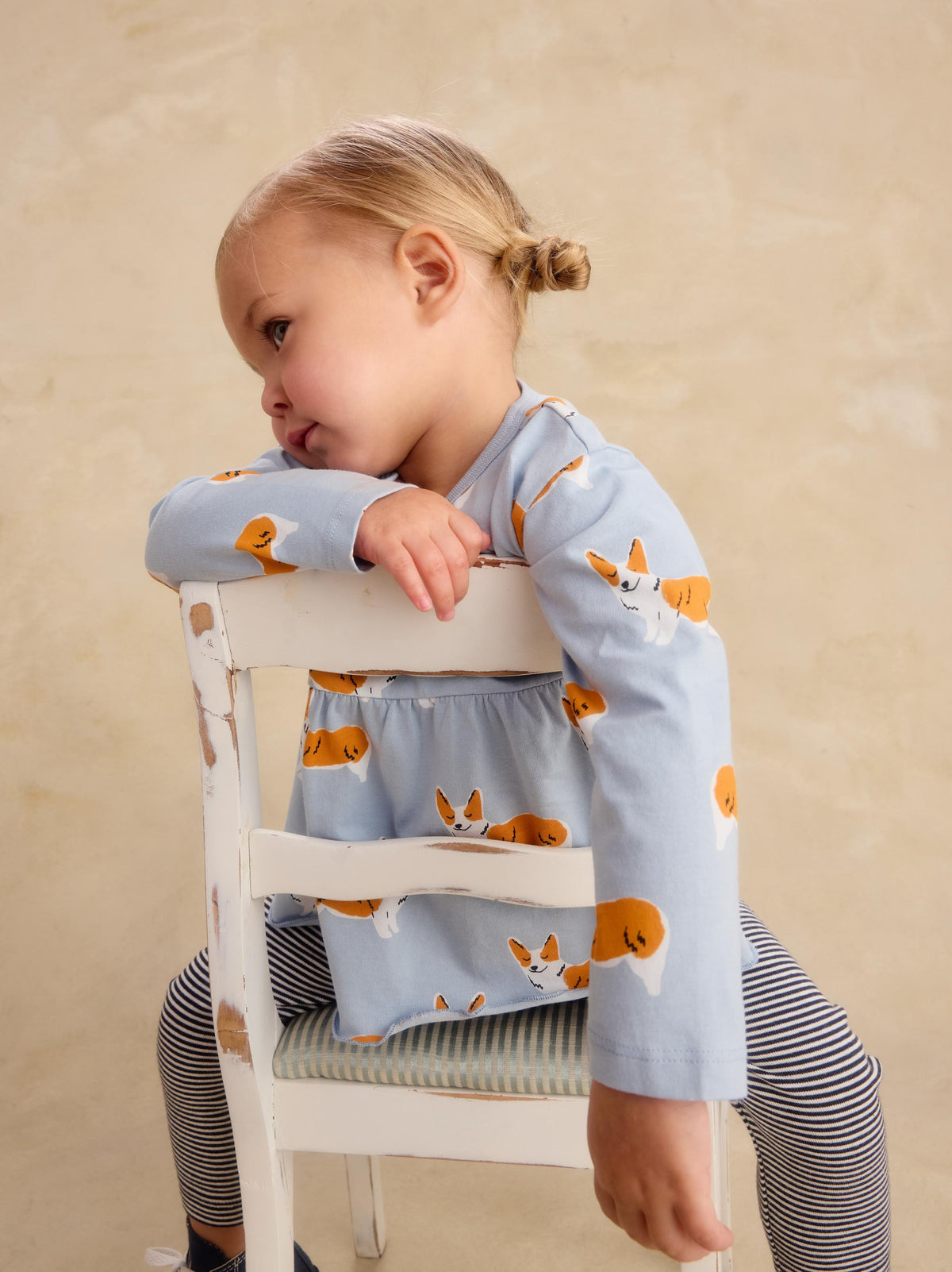 A young child wearing a light blue long-sleeve top and striped leggings sits on a white wooden chair, leaning on the backrest against a warm beige background.
