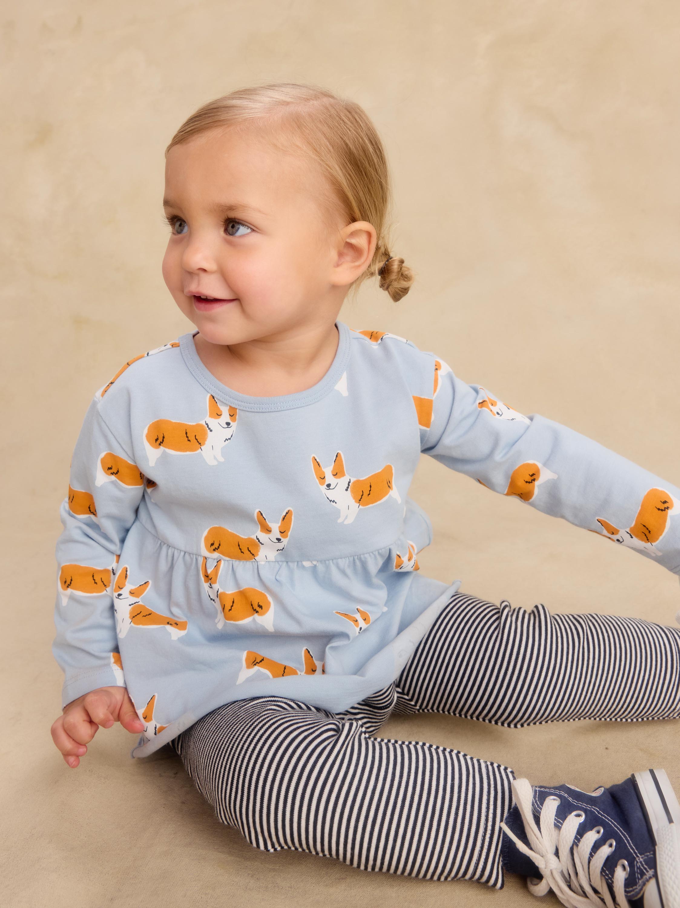 A toddler girl sitting on a beige floor wearing a light blue long-sleeve top with an orange and white corgi dog print, paired with navy and white striped leggings and navy sneakers.
