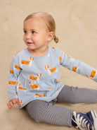 A toddler girl sitting on a beige floor wearing a light blue long-sleeve top with an orange and white corgi dog print, paired with navy and white striped leggings and navy sneakers.