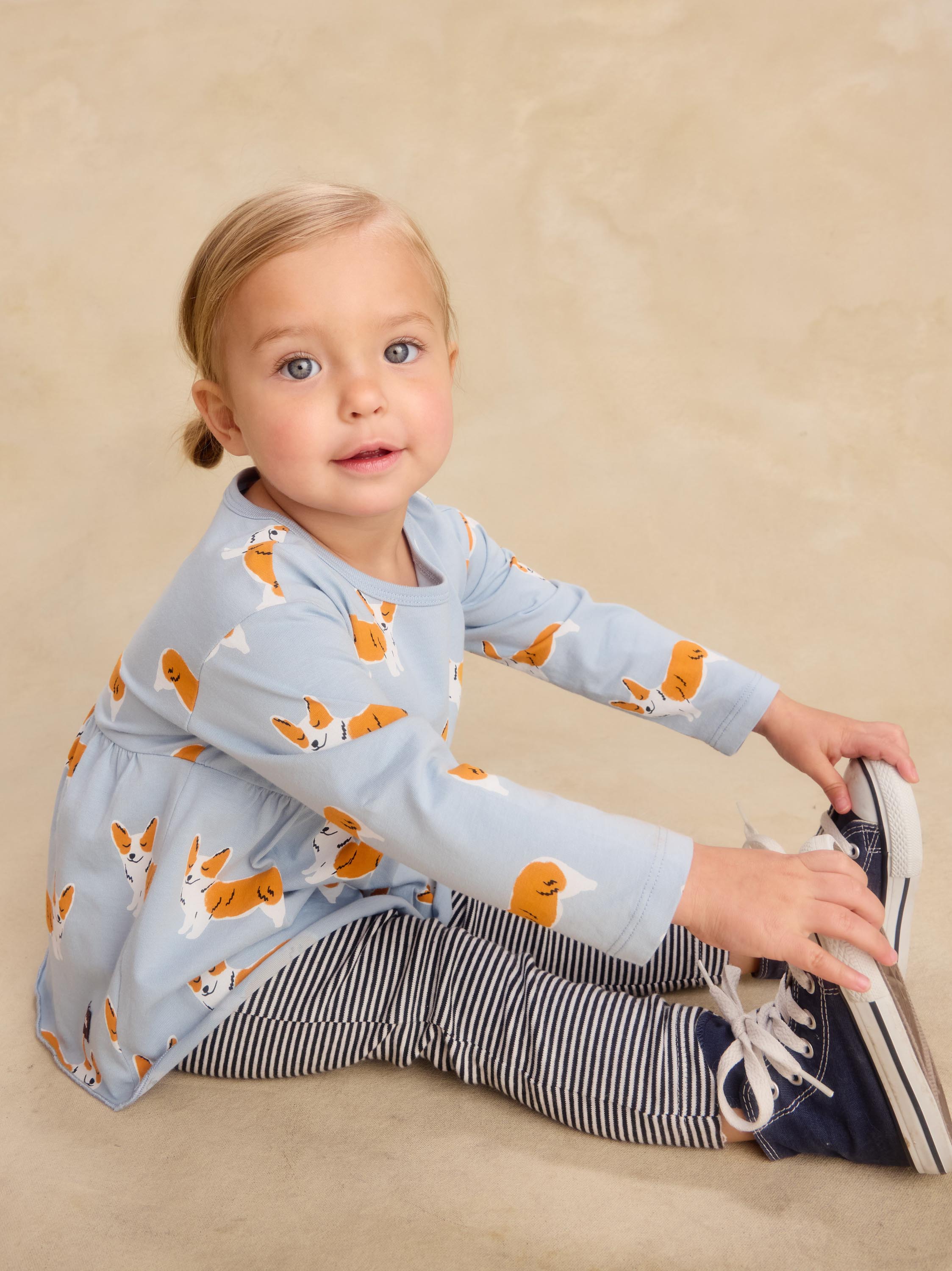 A toddler girl sitting on a beige studio floor wearing a light blue long-sleeve top with an orange and white dog print, navy and white striped leggings, and navy sneakers.