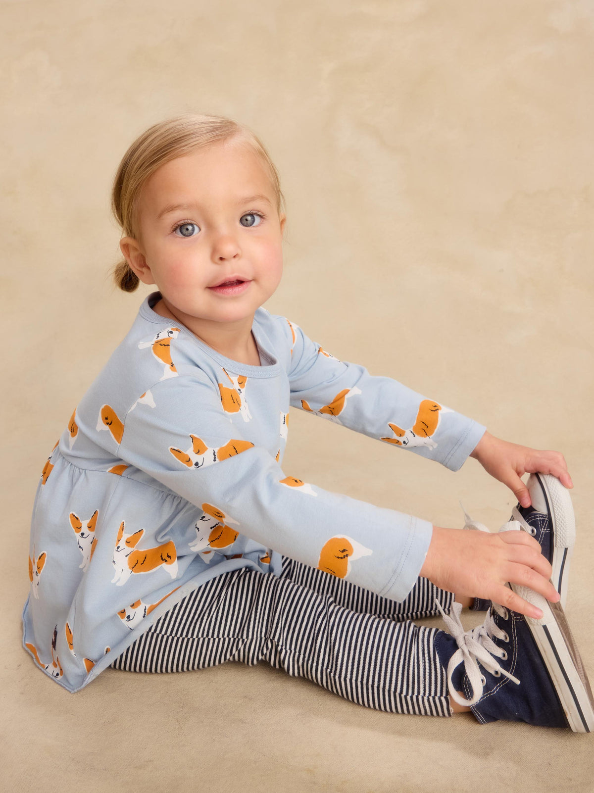 A toddler girl sitting on a beige studio floor wearing a light blue long-sleeve top with an orange and white dog print, navy and white striped leggings, and navy sneakers.