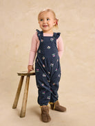 A toddler girl stands next to a small wooden stool against a textured beige backdrop, wearing navy blue corduroy overall with a small floral print, a light pink long-sleeve shirt, and brown boots.