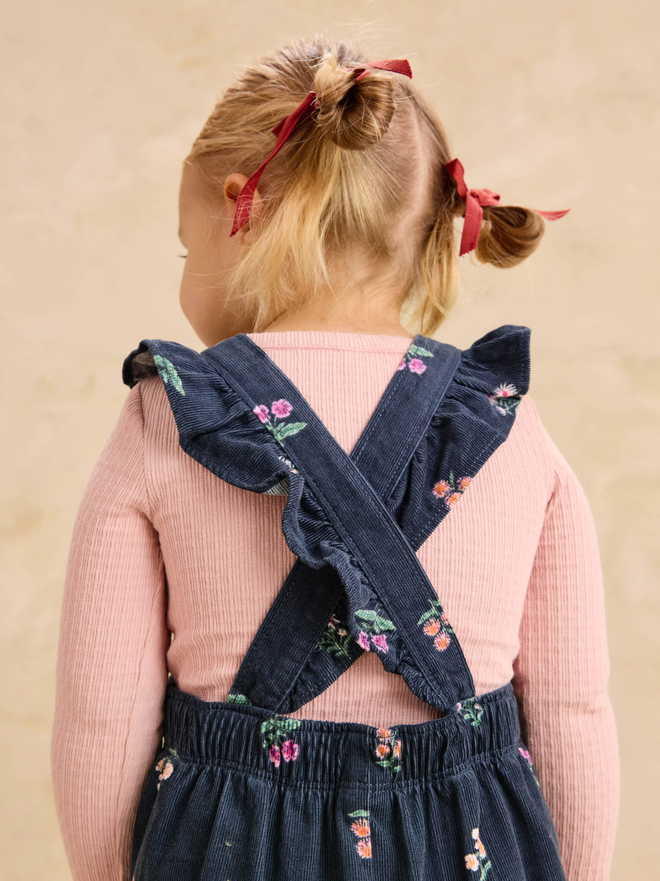 A young child viewed from behind wearing a pink ribbed long-sleeve top and dark navy floral overalls with ruffled shoulder straps, set against a soft beige textured background.