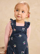 A young toddler girl with blonde hair in a side braid wearing a pink ribbed long-sleeve shirt and a dark navy corduroy overall dress with colorful floral embroidery, posed against a warm beige textured background.