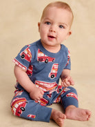 A baby sitting on a beige studio floor wearing a blue short-sleeve romper with a red and white bus print.