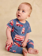 A baby sitting on a beige studio floor wearing a blue short-sleeve romper and matching pants with a red double-decker bus pattern.