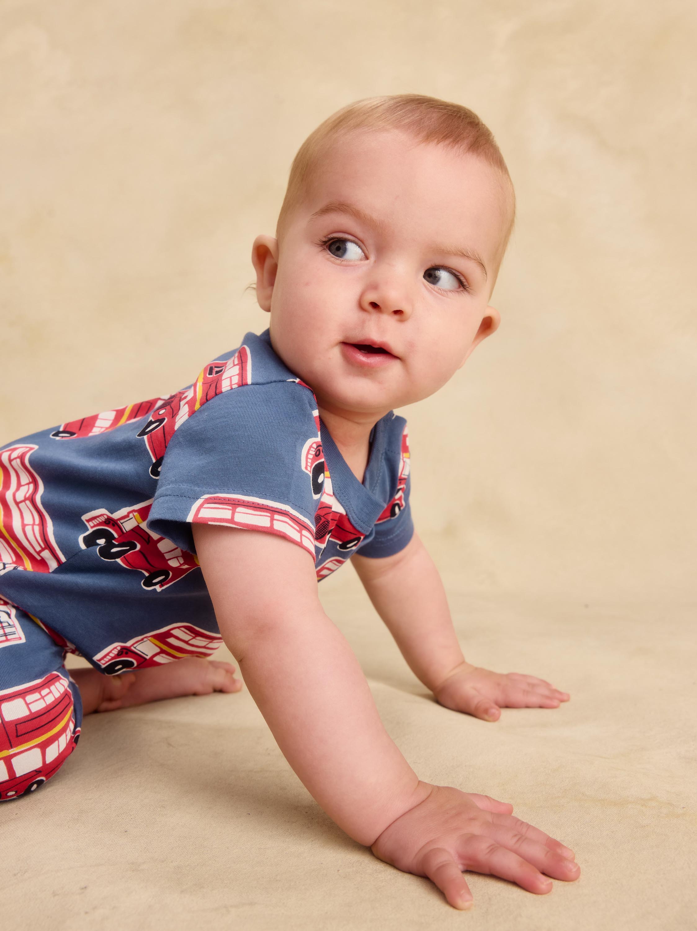 A baby crawling on a beige studio backdrop, wearing a short-sleeve blue romper with a red and white fire truck pattern.