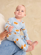 A baby wearing a light blue long-sleeve sleeper with an orange and white dog print, sitting on an adult's lap against a beige textured background.