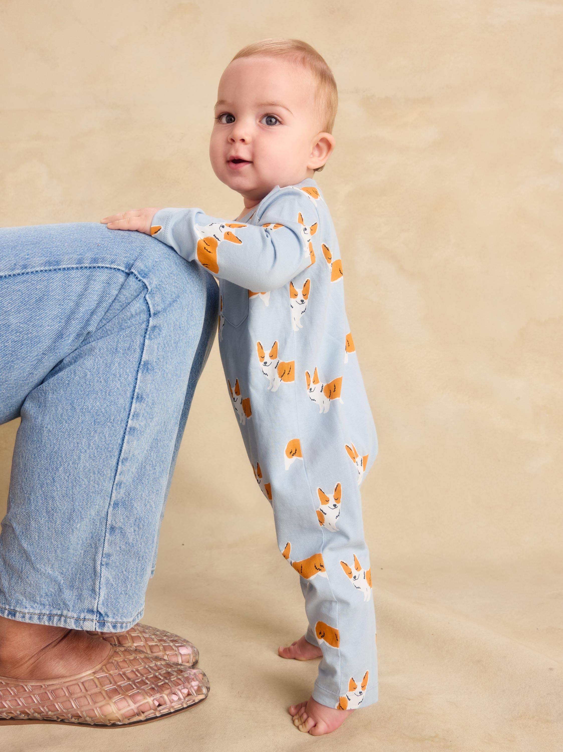 A baby wearing a light blue long-sleeve footless jumpsuit with an orange and white corgi print, standing next to an adult wearing blue jeans against a beige background.