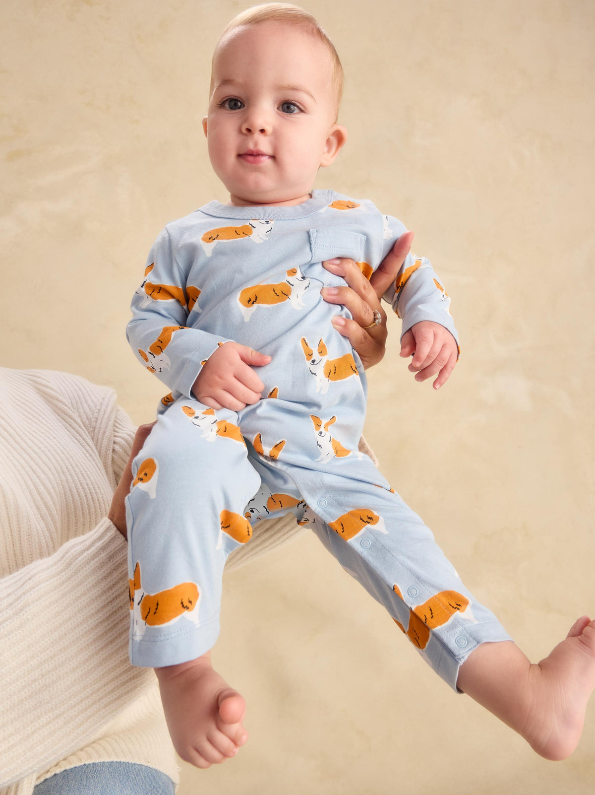A baby sitting on a cream ribbed cushion, wearing a light blue long-sleeve pajama set with an orange and white dog print. The baby is being held by an adult whose hands are visible.