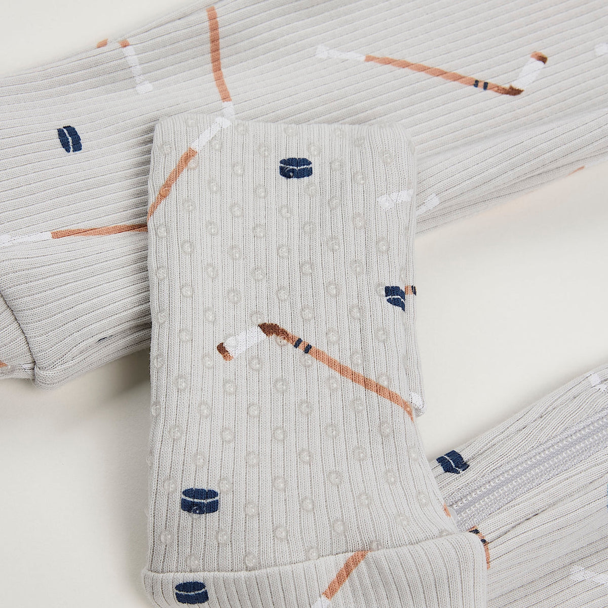 Close-up a light gray ribbed fabric, featuring an all-over print of hockey pucks and sticks in brown, white, and navy blue on a white backgroud.