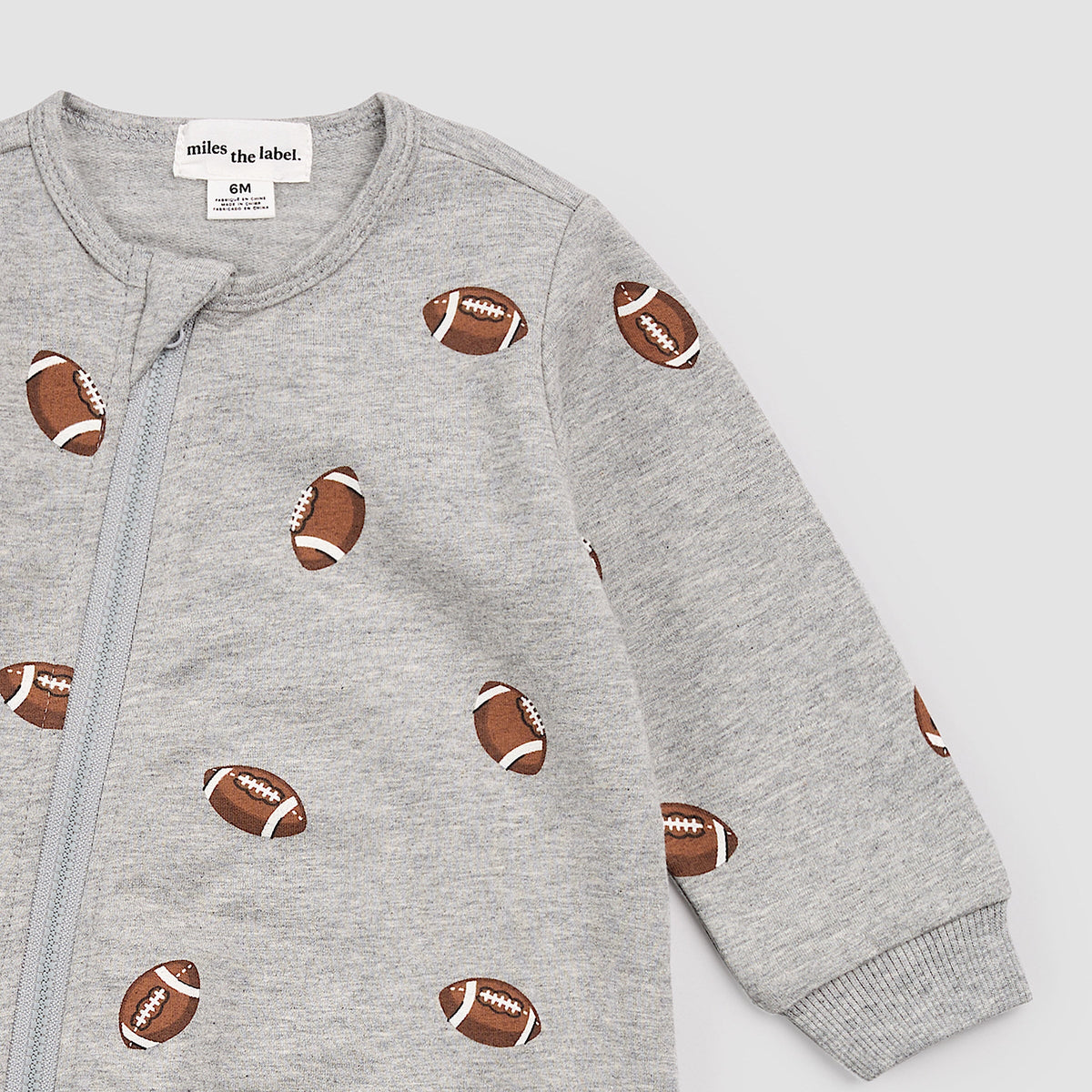 Close-up a baby long-sleeve footless one-piece sleeper in light heather gray, featuring an all-over print of brown and white footballs and a front zipper.