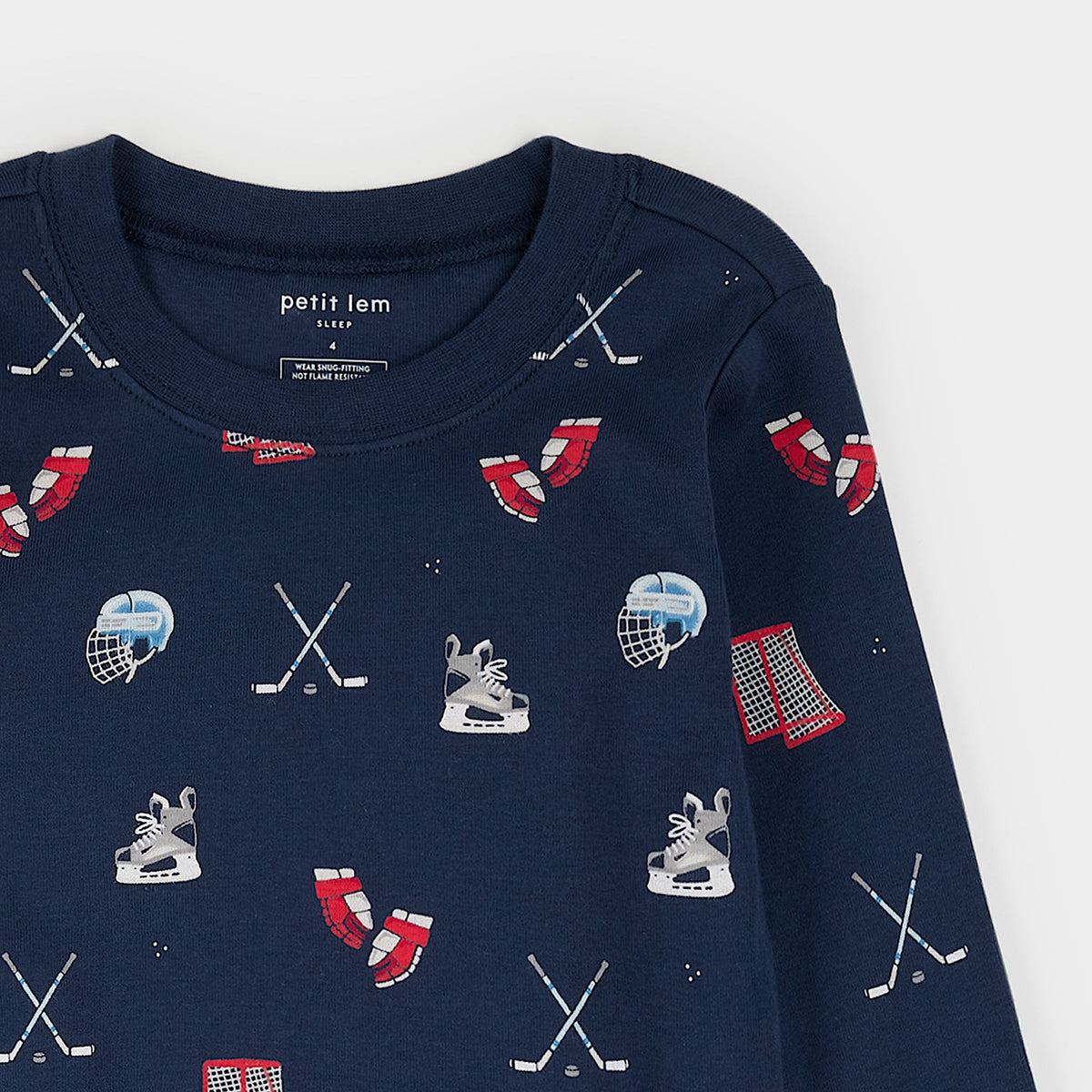 A close-up of a navy blue long-sleeve children's top with an all-over hockey-themed print, including crossed hockey sticks, skates, helmets, and red-and-white socks. The inner neck label reads 'petit lem'.