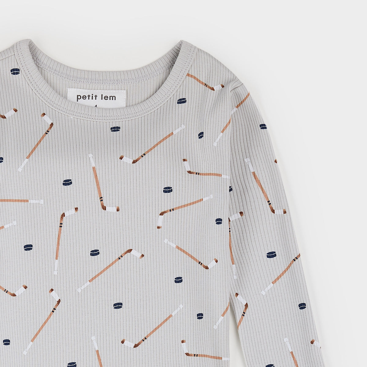 A close-up of a child's long-sleeve ribbed knit top in light gray, featuring an all-over pattern of hockey sticks and pucks in brown, white, and navy blue. The brand label 'petit lem' is visible inside the collar.