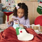 A young girl in a purple sweater sits under a red knit blanket, looking into a white Christmas stocking. The stocking has a green scarf and red and blue pom-poms, and she holds a package of Christmas cookies.