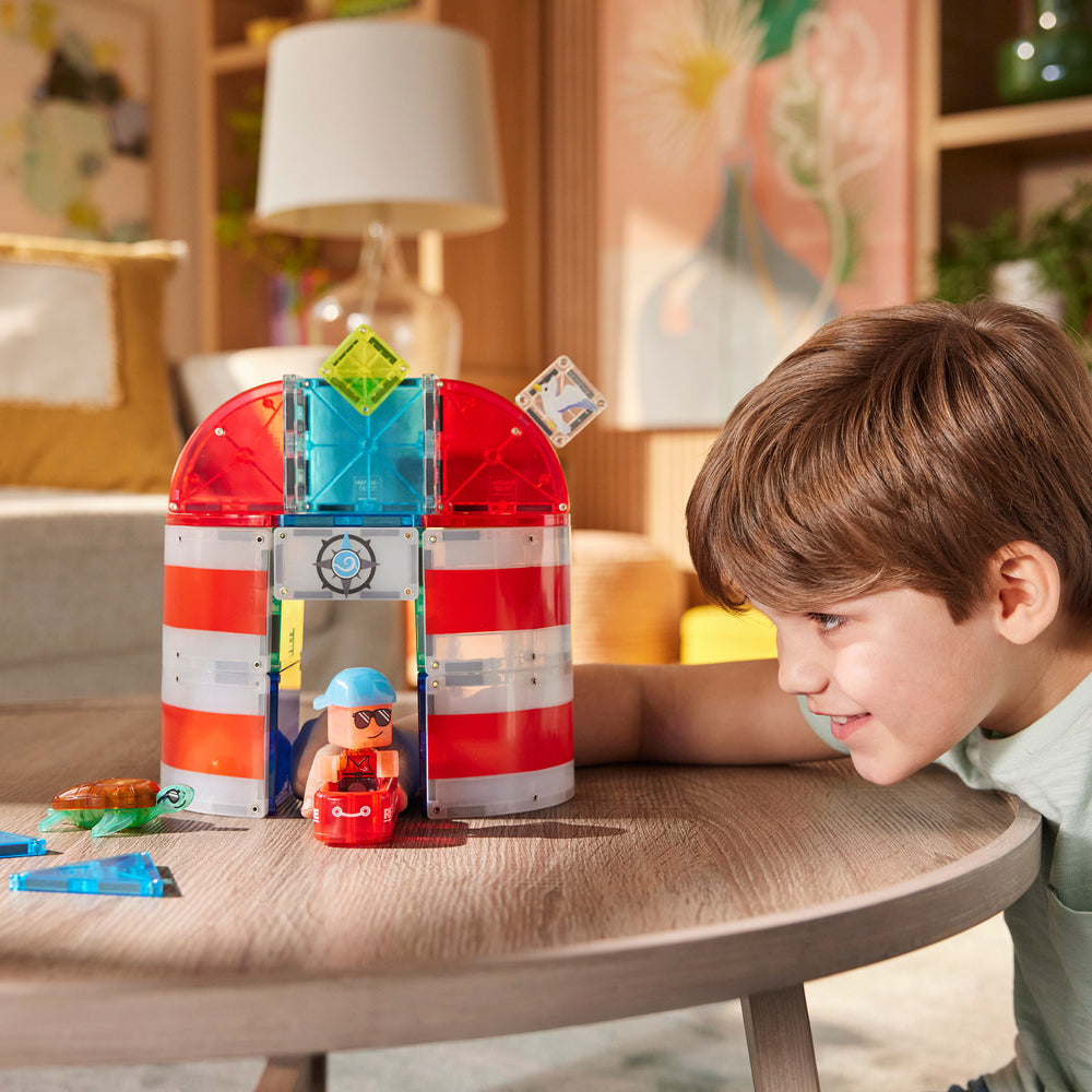 A young boy smiling as he plays with the Magna-Tiles Lighthouse Rescue set on a coffee table, building a red-and-white striped lighthouse structure with a rescue raft and sea turtle figurine nearby.