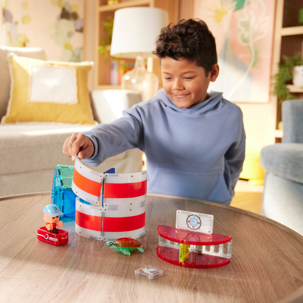 A young boy smiling as he plays with the Magna-Tiles Lighthouse Rescue set on a coffee table, building a red-and-white striped lighthouse structure with a rescue raft and sea turtle figurine nearby.