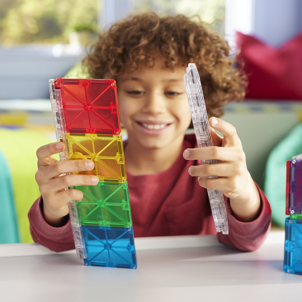 A smiling child holds colorful magnetic tiles and a clear rail piece from the Magna-Tiles Rail Racers set, playing at a table.