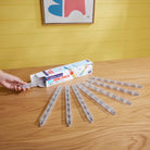 An open box of the Magna-Tiles Rail Racers Rail Pack rests on a wooden table, with eight clear rail pieces fanned out alongside it and a hand holding one piece.