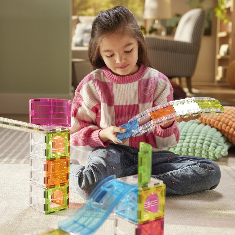 A young girl sits on a carpeted floor in a living room, playing with translucent magnetic building blocks. She is wearing a pink and white checkered sweater and blue jeans, holding a blue curved magnetic piece.