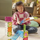 A young girl sits on a carpeted floor in a living room, playing with translucent magnetic building blocks. She is wearing a pink and white checkered sweater and blue jeans, holding a blue curved magnetic piece.