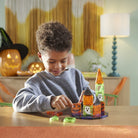 A young boy wearing a gray sweatshirt sits at a wooden table, playing with a colorful magnetic building toy castle made of translucent orange, green, and black pieces. The background shows a cozy, softly lit room with Halloween decorations, including a jack-o'-lantern and a witch hat on a shelf, and a blue-green spiral lamp on a table lamp stand.