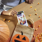 Person holding a smartphone with a Halloween-themed app over a burlap bag with a pumpkin design, surrounded by candy corn on a wooden table.