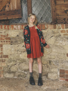 A young girl standing in front of a weathered stone wall, wearing a long dark knit cardigan with a red-and-green floral pattern over a reddish-orange dress, paired with dark boots and socks.