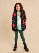 A young girl standing against a neutral backdrop, wearing a dark knit long cardigan with a bold red floral pattern, a light green graphic t-shirt, green pants, and dark boots.