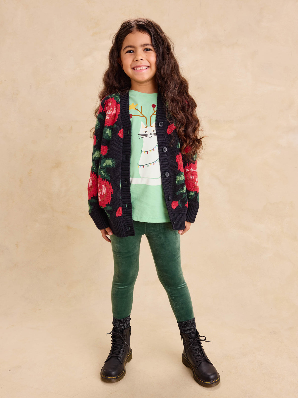 A young girl standing against a neutral backdrop, wearing a dark knit long cardigan with a bold red floral pattern, a light green graphic t-shirt, green pants, and dark boots.