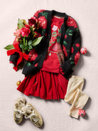 Flat lay of a dark knit cardigan with a red-and-green floral pattern, paired with a bright red skirt and top, a bouquet of red flowers with greenery, cream-colored tights, and gold shoes.