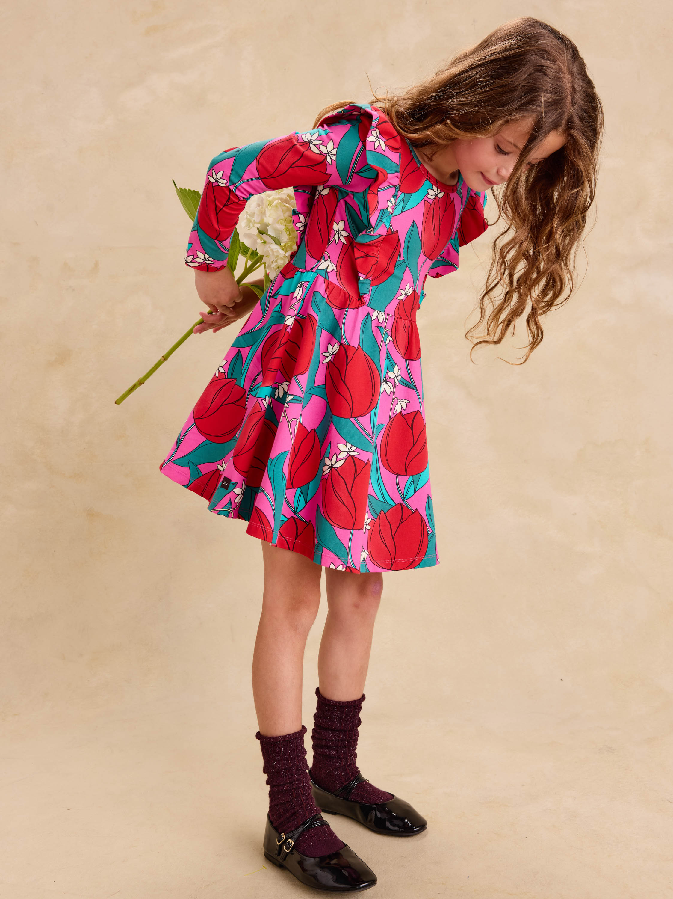 Ruffle Shoulder Ballet Dress - Tulip Twist in Pink by Tea Collection featuring long sleeves, a crew neckline, ruffled shoulder details, and an artist-drawn red and teal tulip floral pattern over a pink background, worn with dark socks and shoes while holding a flower.