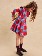 Ruffle Shoulder Ballet Dress - Tulip Twist in Pink by Tea Collection featuring long sleeves, a crew neckline, ruffled shoulder details, and an artist-drawn red and teal tulip floral pattern over a pink background, worn with dark socks and shoes while holding a flower.