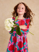 Ruffle Shoulder Ballet Dress - Tulip Twist in Pink by Tea Collection featuring long sleeves, a crew neckline, ruffled shoulder details, and an artist-drawn red and teal tulip floral pattern over a pink background, worn by a smiling child holding a large white flower.