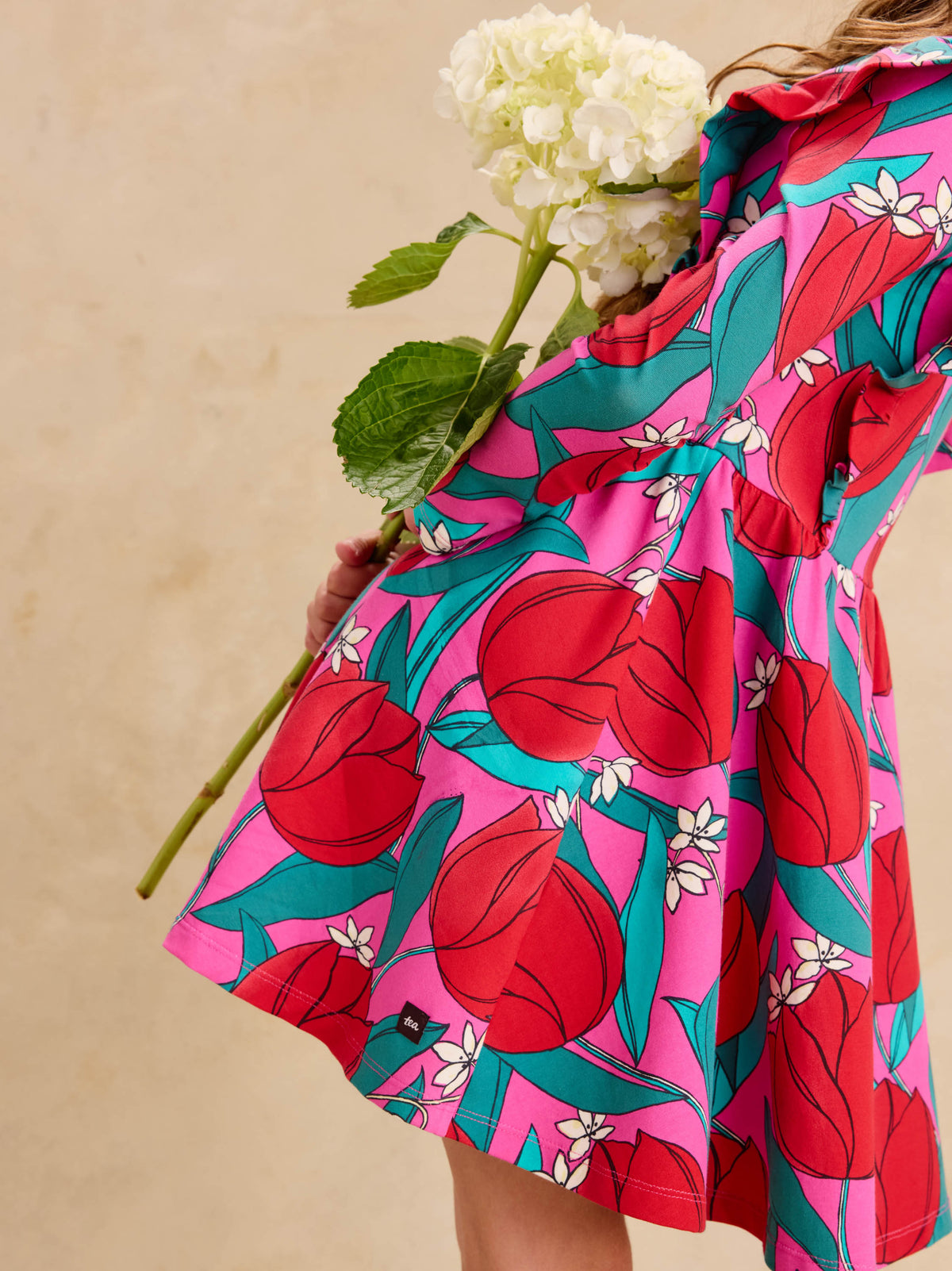 Ruffle Shoulder Ballet Dress - Tulip Twist in Pink by Tea Collection featuring long sleeves, a crew neckline, ruffled shoulder details, and an artist-drawn red and teal tulip floral pattern over a pink background, viewed from the side while holding a large white flower.