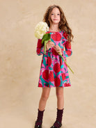 Ruffle Shoulder Ballet Dress - Tulip Twist in Pink by Tea Collection featuring long sleeves, a crew neckline, ruffled shoulder details, and an artist-drawn red and teal tulip floral pattern over a pink background, worn by a child standing while holding a large white flower stem against a neutral background.