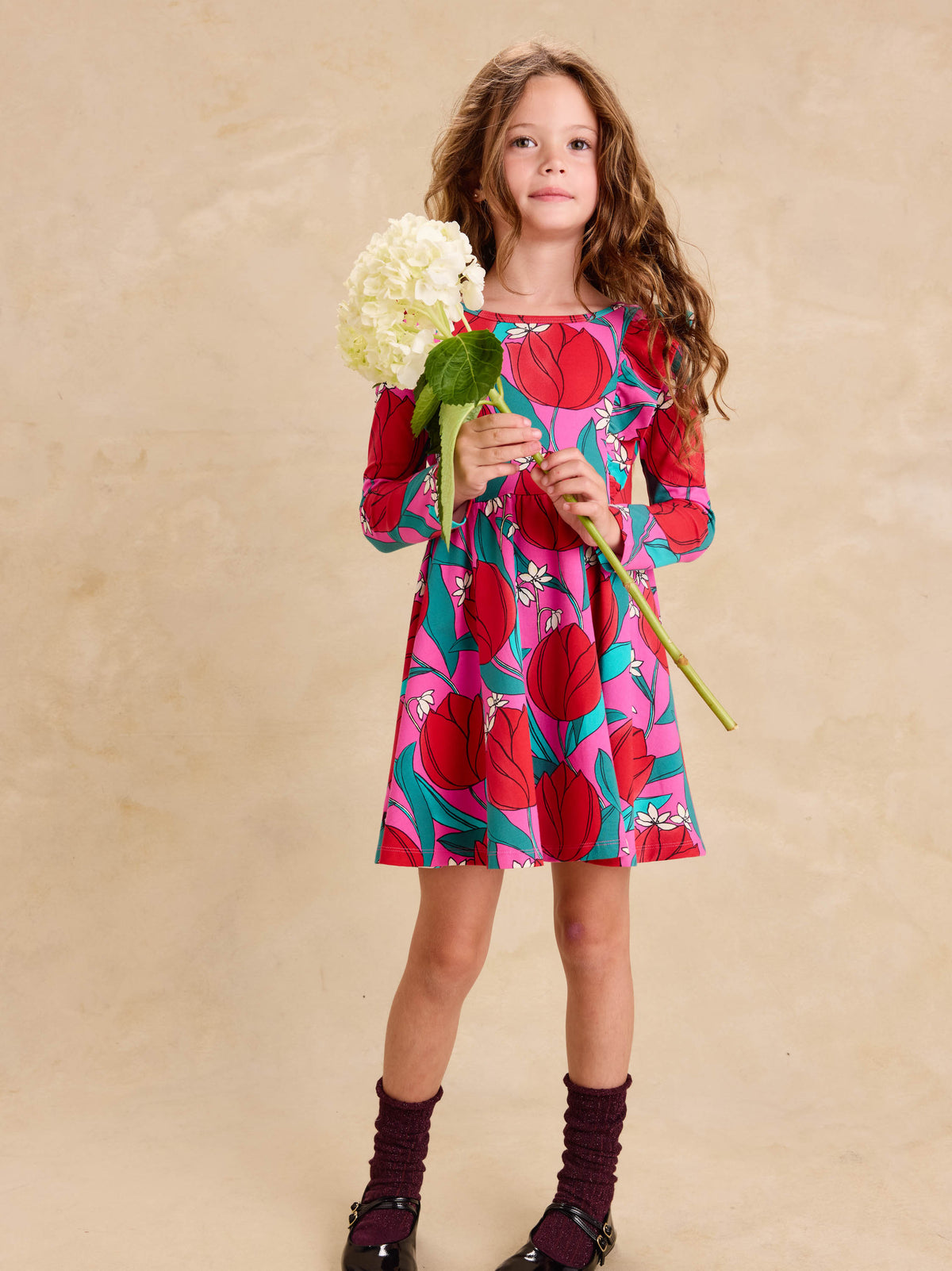 Ruffle Shoulder Ballet Dress - Tulip Twist in Pink by Tea Collection featuring long sleeves, a crew neckline, ruffled shoulder details, and an artist-drawn red and teal tulip floral pattern over a pink background, worn by a child standing while holding a large white flower stem against a neutral background.