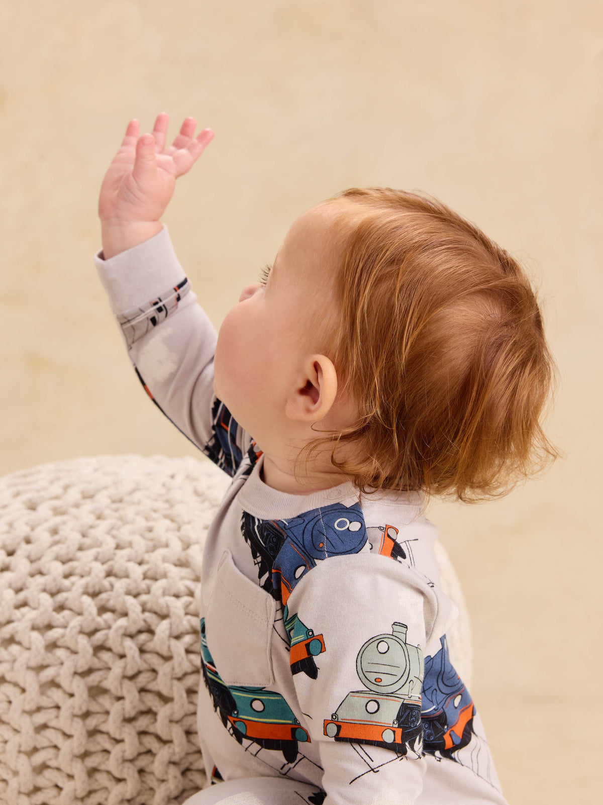 Baby wearing a long sleeve romper with a vintage steam locomotive train print, reaching upward with one hand.