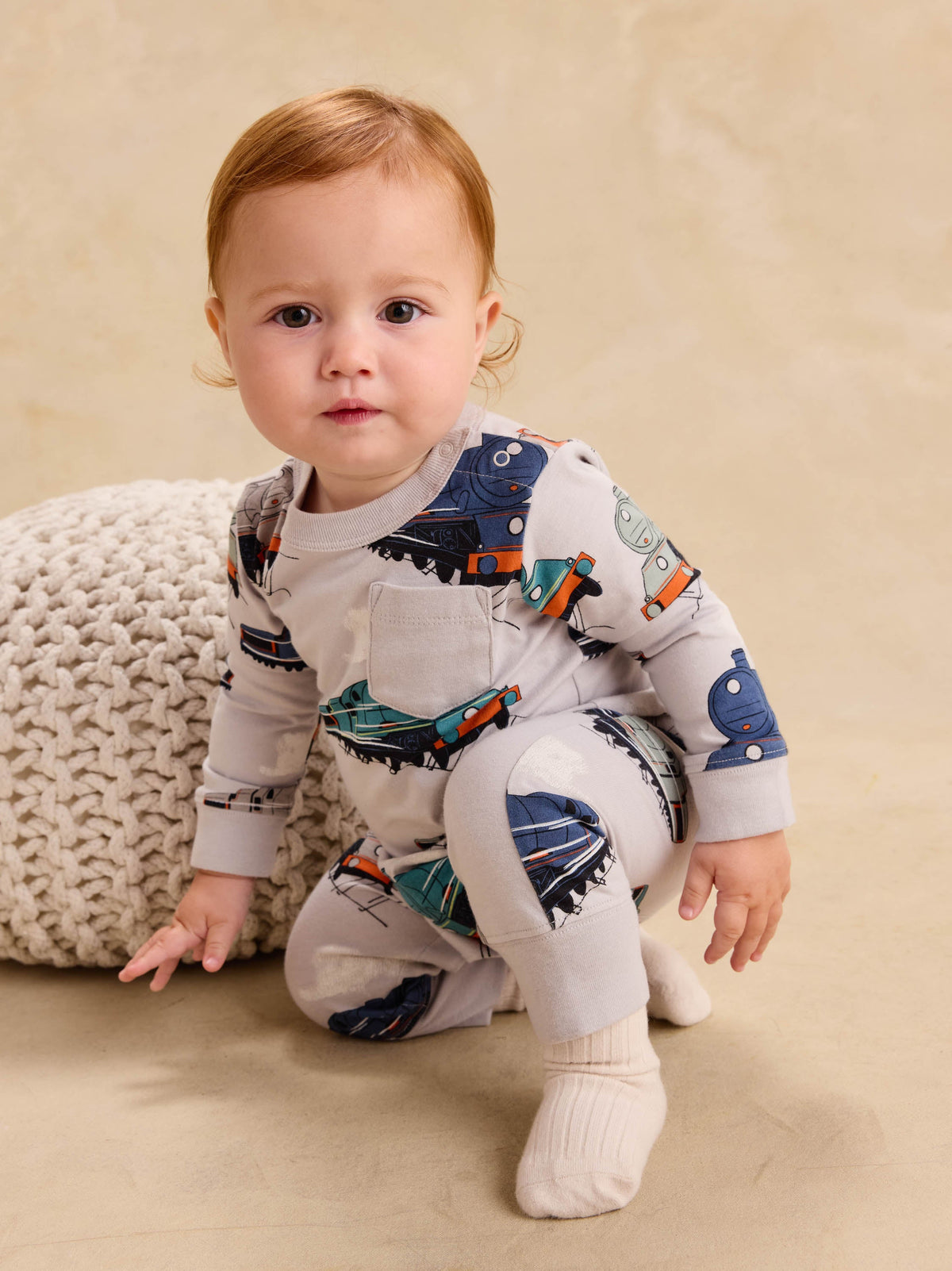 Baby wearing a long sleeve romper with a vintage steam locomotive train print, sitting next to a knit cushion.