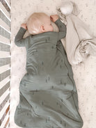 A baby sleeping soundly in a swaddle blanket inside a crib. The swaddle is sage green with a small tree pattern, and the baby is lying on a light cream floral mattress with a beige blanket nearby.