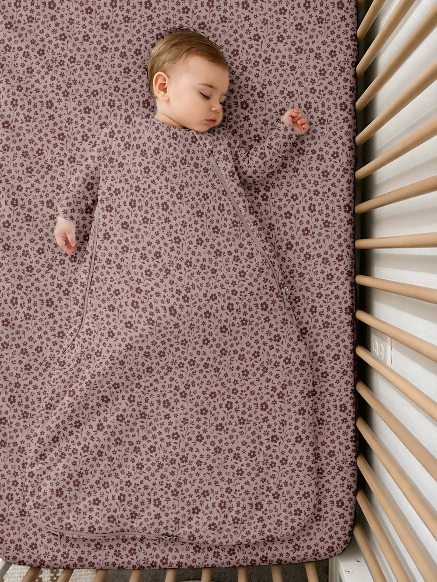 A sleeping baby wearing a long-sleeve sleep sack with a small floral pattern, lying on a mattress inside a wooden crib.