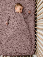 A sleeping baby wearing a long-sleeve sleep sack with a small floral pattern, lying on a mattress inside a wooden crib.