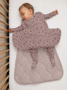 A sleeping baby wearing a dusty mauve footed sleeper with a small floral pattern, lying on a white mattress next to a wooden crib.