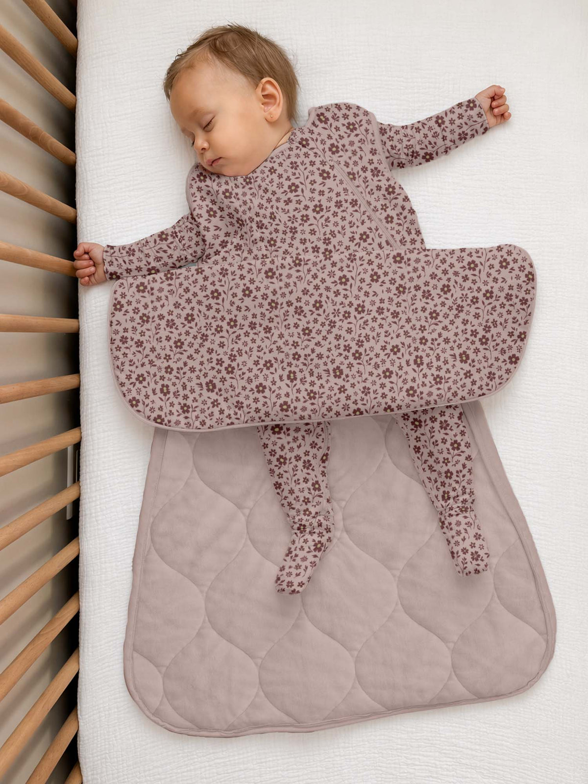 A sleeping baby wearing a dusty mauve footed sleeper with a small floral pattern, lying on a white mattress next to a wooden crib.