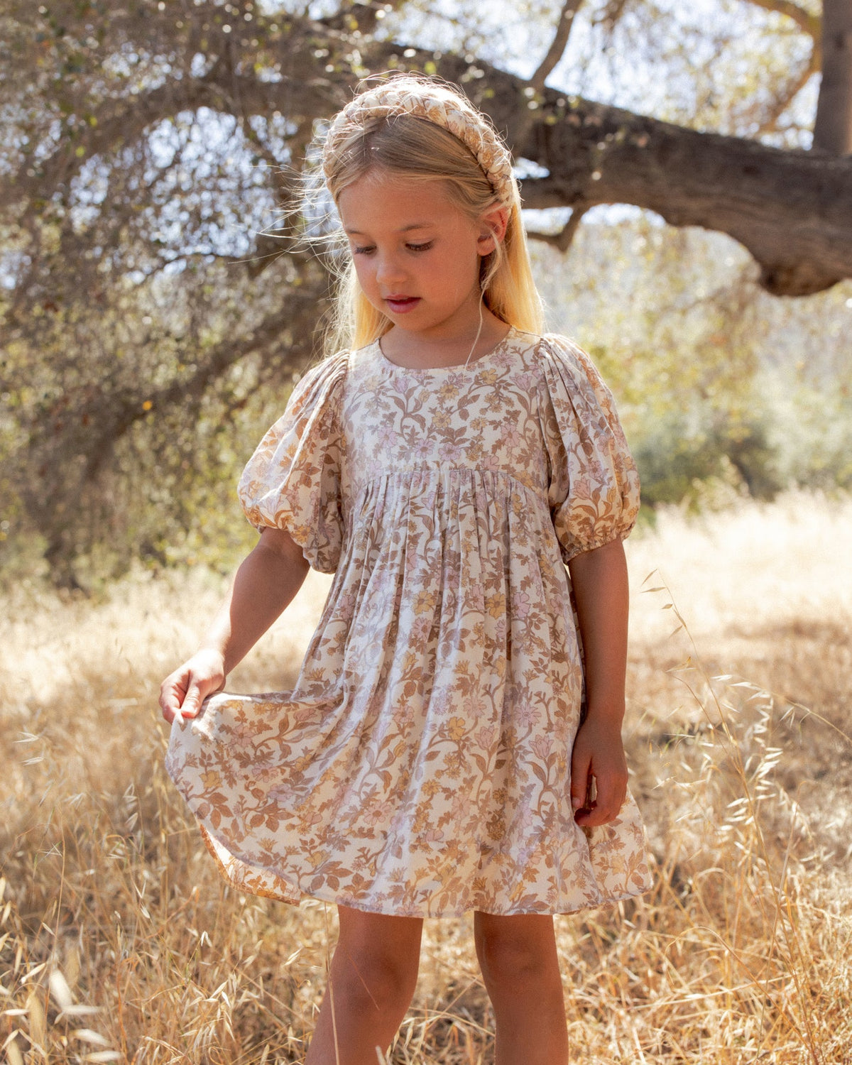 A young girl with blonde hair in a headband stands in a dry grassy outdoor setting wearing a short-sleeved, knee-length dress with a beige and brown floral pattern.